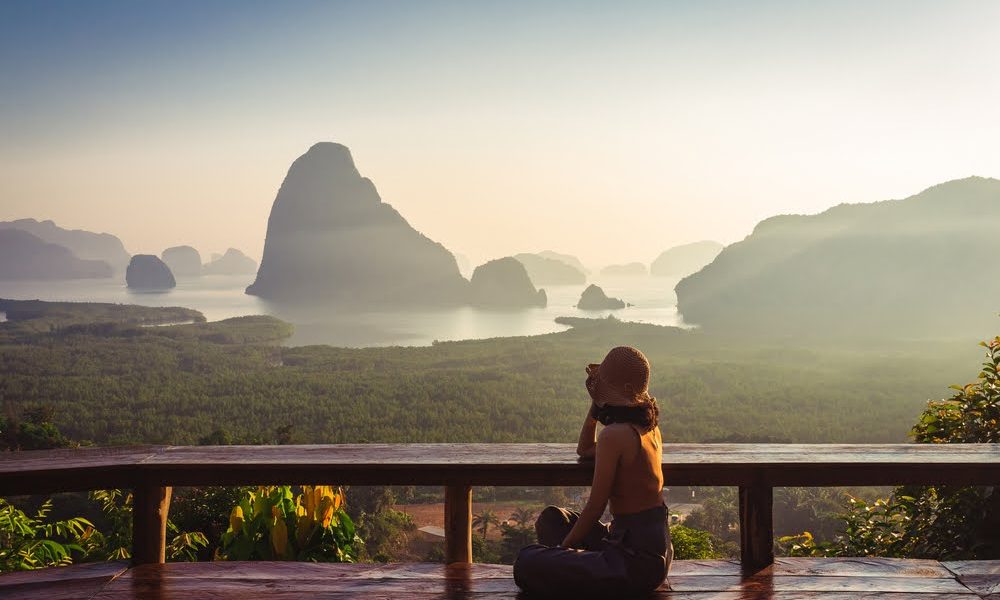 Solo woman traveler is relaxing on balcony with aerial view of islands and mangrove forest in the early morning, Phang nga, Thailand. Travel lifestyle and healthy environment concepts in soft tone.